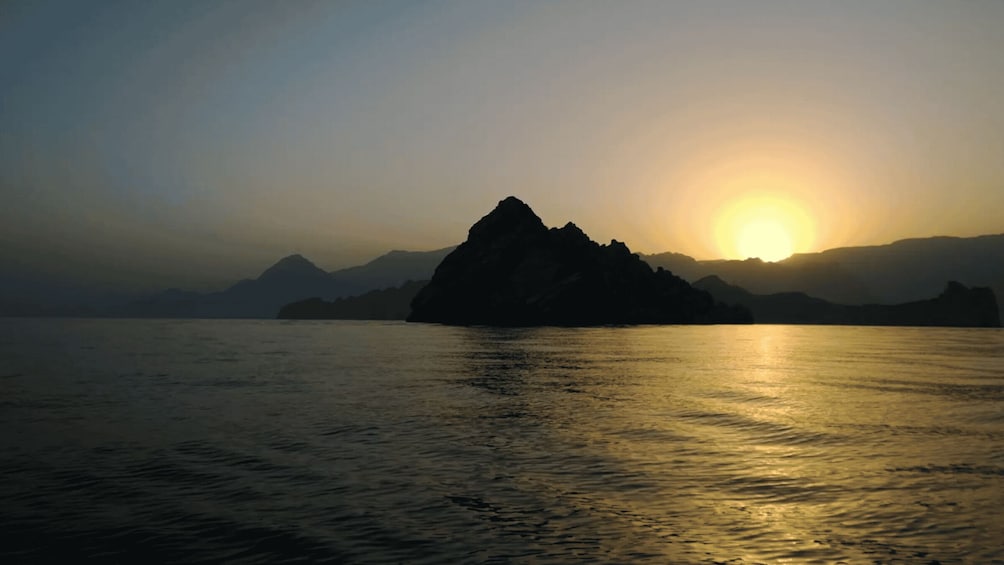 A sunset over a calm body of water with a prominent rock formation in the foreground and mountains in the background. 2