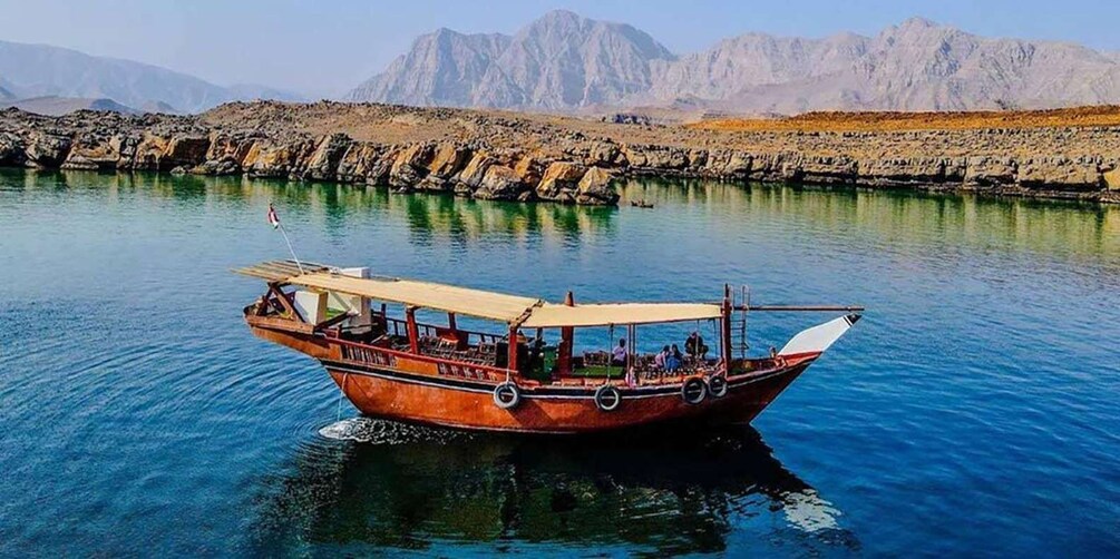 A wooden boat with passengers on a calm body of water, surrounded by rocky terrain and mountains in the background. 1