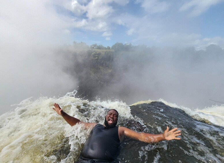 A person in a black wetsuit with arms outstretched in a turbulent water body, possibly a river or a small waterfall, with mist and greenery in the background. 3