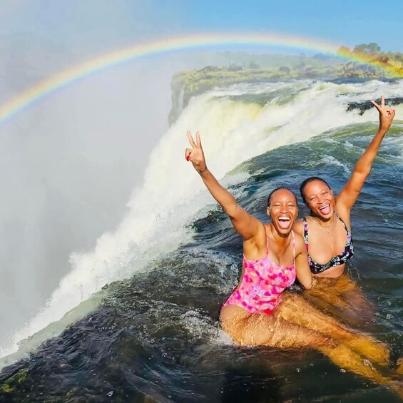 Two people in a body of water with a waterfall and a rainbow in the background. 2