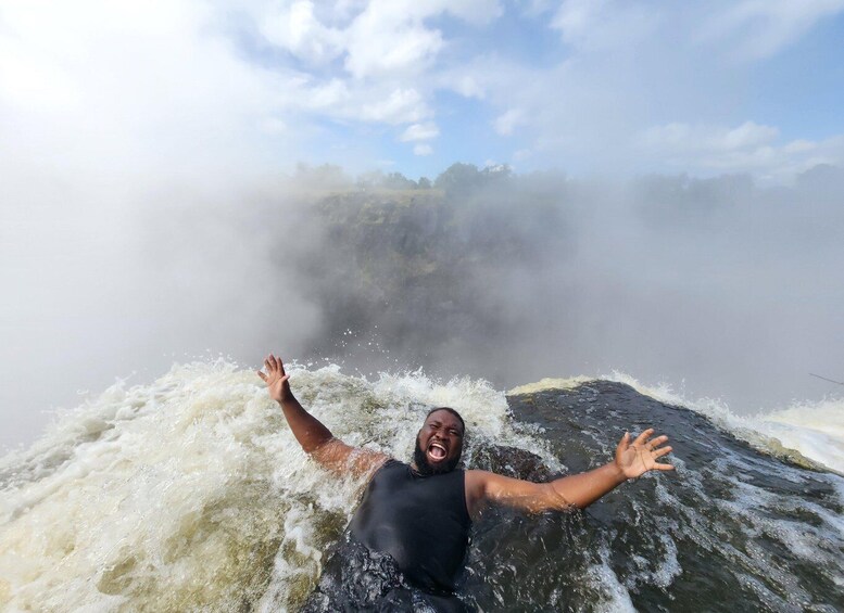 A person in a black wetsuit with arms outstretched in a turbulent river surrounded by mist and rocks. 1