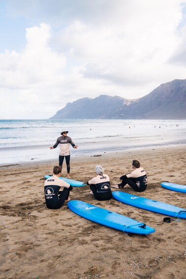 A beach with surfers sitting on the sand, a surf instructor standing, and blue surfboards. 3