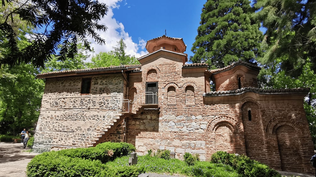 A historic stone building with a bell tower, surrounded by lush greenery and trees. 2