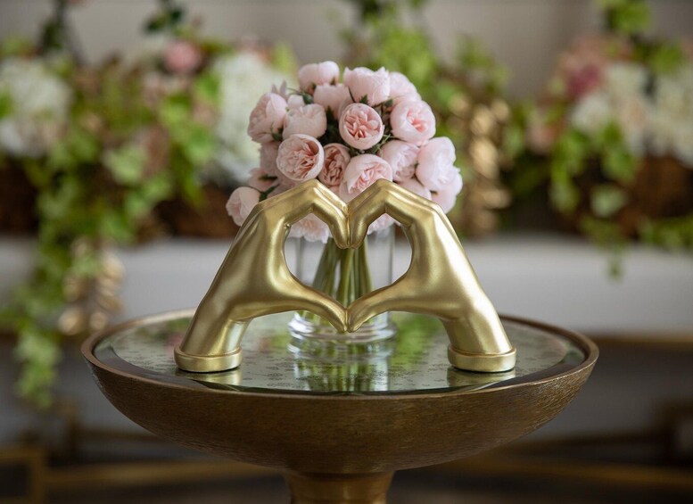 A golden heart-shaped object on a table with a vase of pink flowers in the background. 5