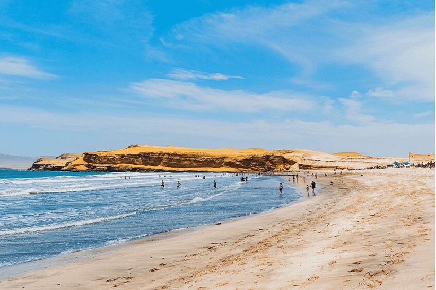 A beach with people swimming and walking along the shore, with a large rock formation in the background. 2