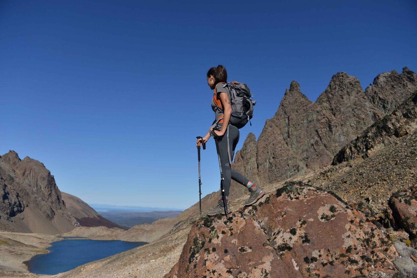 A hiker on a rocky mountain peak with a backpack, holding hiking poles, and a clear blue sky in the background. 1