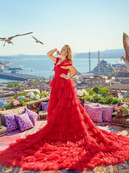 A person in a red dress standing on a balcony with a view of a cityscape, including a mosque and a body of water. 3
