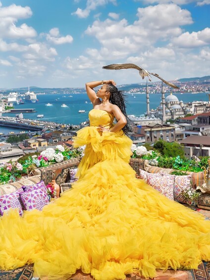 A person in a yellow dress on a balcony with a cityscape and sea in the background. 1