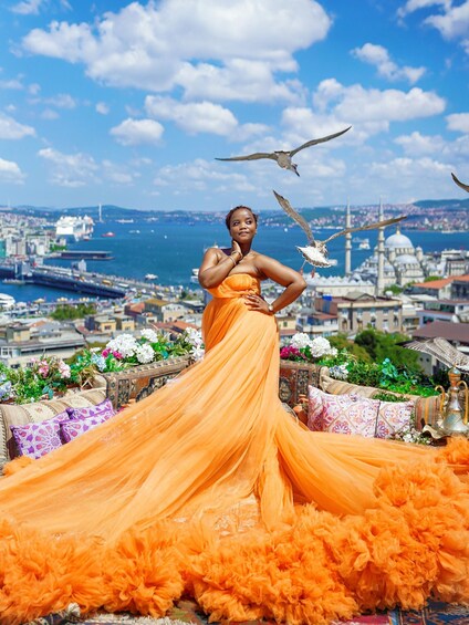 A person in an orange dress standing on a balcony with a cityscape and sea in the background. 2