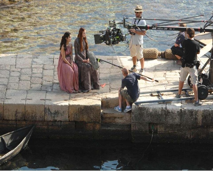 A film crew on a dock by water, with actors in period costumes, a camera, and equipment. 1