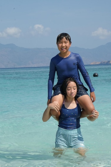 A person riding another on shoulders in clear turquoise water with mountains in the background. 2