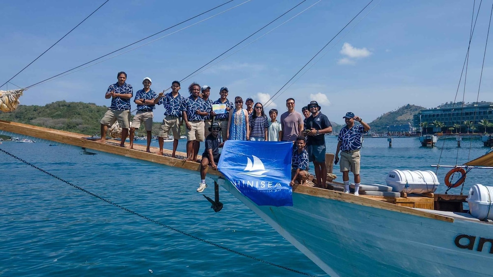 A group of people on a wooden boat with a blue sail, anchored in a body of water with a distant shoreline and buildings. 4