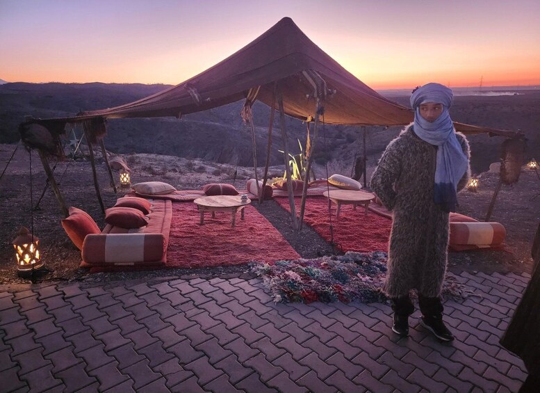 A desert campsite with a large tent, red cushions, and lanterns at sunset. 3
