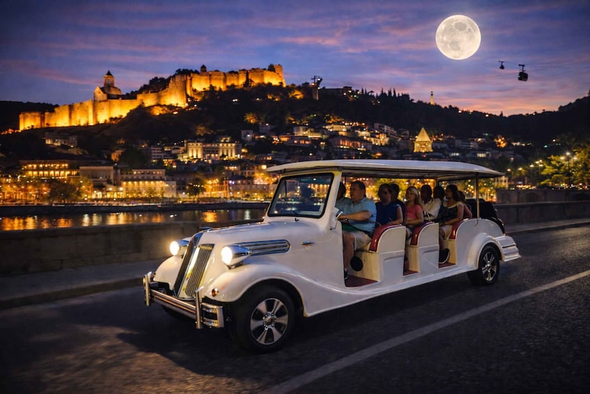 A vintage car with passengers driving past a historic castle at night. 1