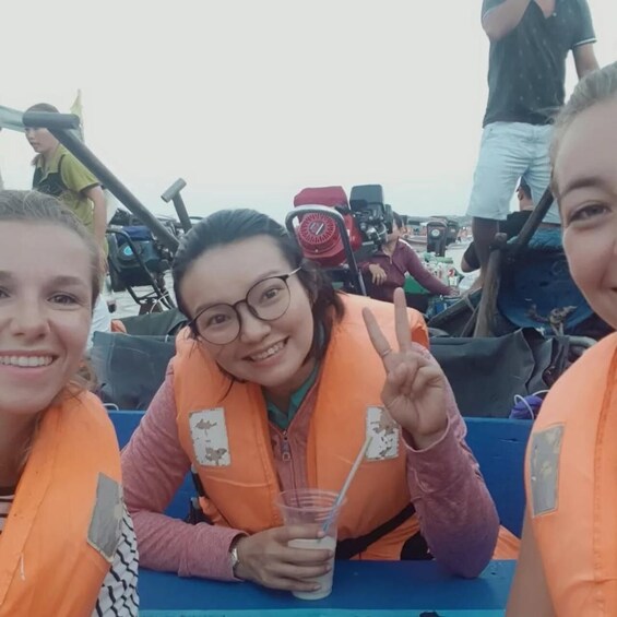 Three individuals in orange life jackets, one holding a drink, on a boat with other people and equipment in the background. 2