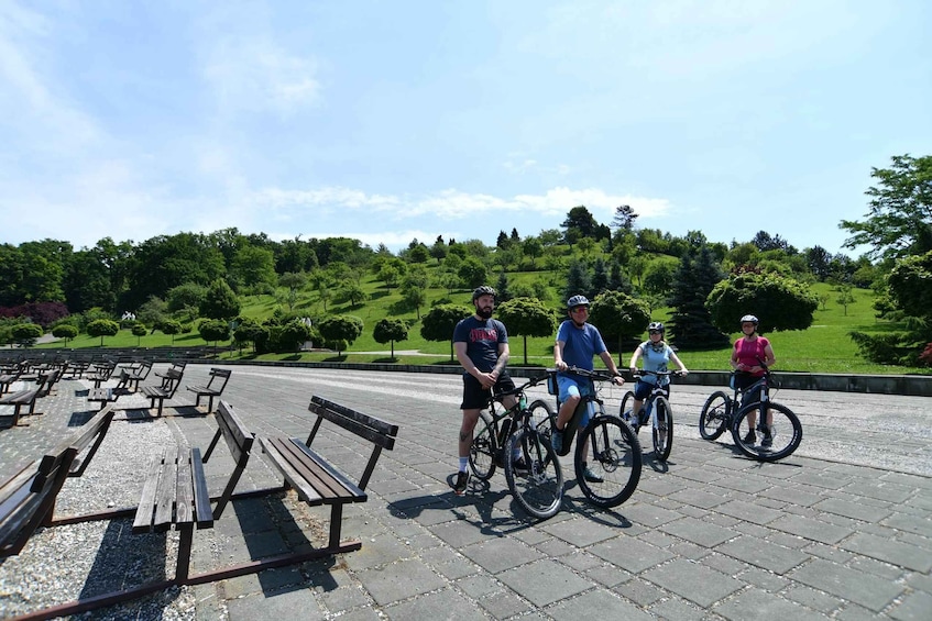 Four people on bicycles in a park with benches and green hills in the background. 1