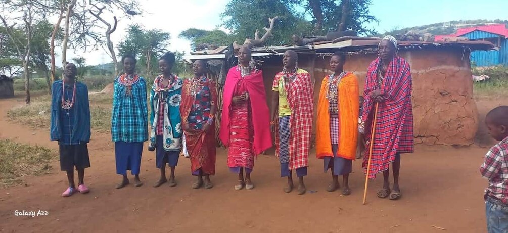 A group of people in traditional attire standing in a rural setting with a dirt path and a simple structure in the background. 4