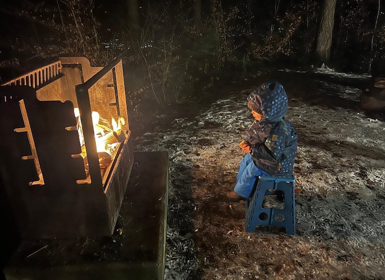 A person sitting on a stool next to a fire pit in a wooded area. 4