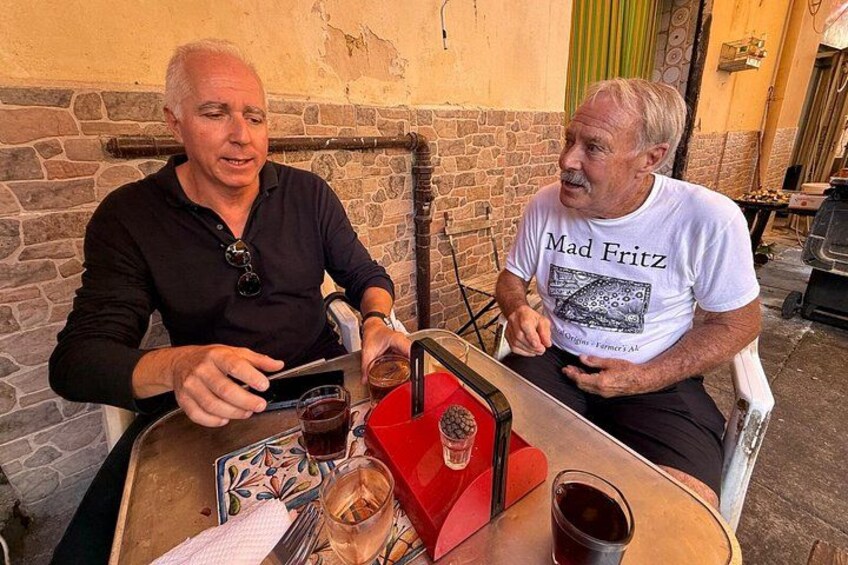 Två män sitter vid ett bord utomhus med drycker och en röd låda, som eventuellt innehåller mat eller n ågot att dricka. 1