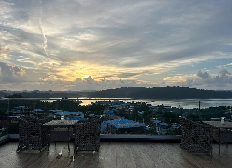 A sunset view from a rooftop with wicker furniture, overlooking a coastal town and mountains. 4