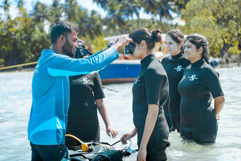A person in a blue shirt taking a photo of two individuals in black wetsuits standing in shallow water with a boat in the background. 5