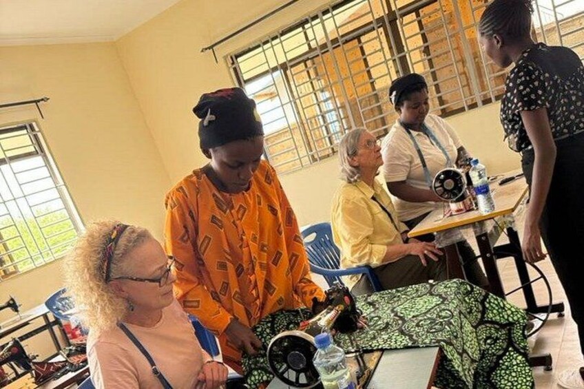 A group of people engaged in a discussion around a table with a sewing machine, in a room with barred windows and a tiled floor. 5