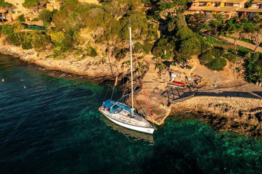 A sailboat anchored near a rocky coastline with a clear blue sea and a small village in the background. 1