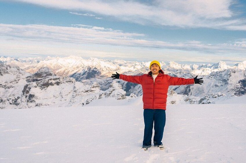 Una persona de pie en la cima nevada de una montaña, con los brazos extendidos, vistiendo una chaqueta roja y un gorro amarillo. 1