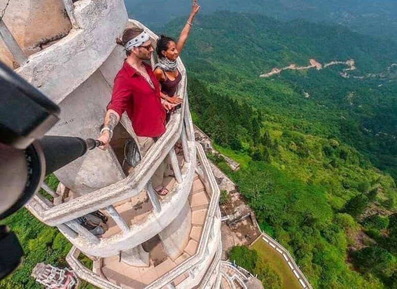 A couple standing on a high observation deck with a view of a lush green valley and winding roads below. 1