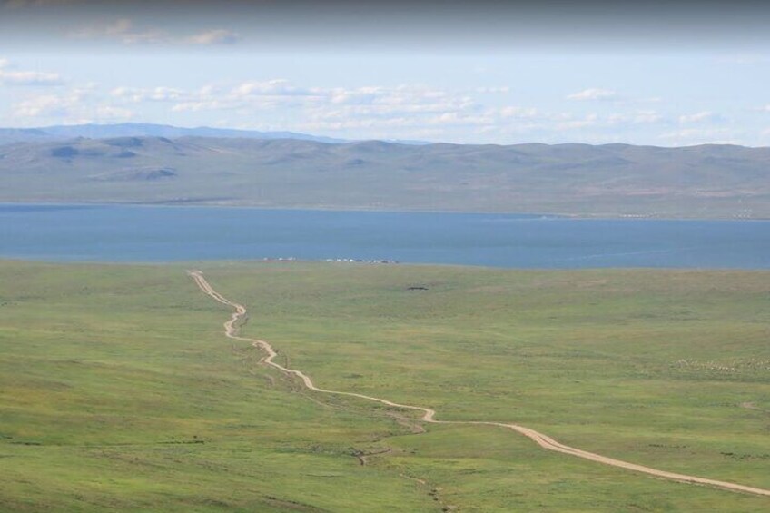 A winding dirt road in a grassy landscape with a body of water and mountains in the background. 3