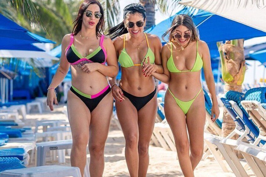 Three people walking on a beach with lounge chairs and umbrellas in the background. 4