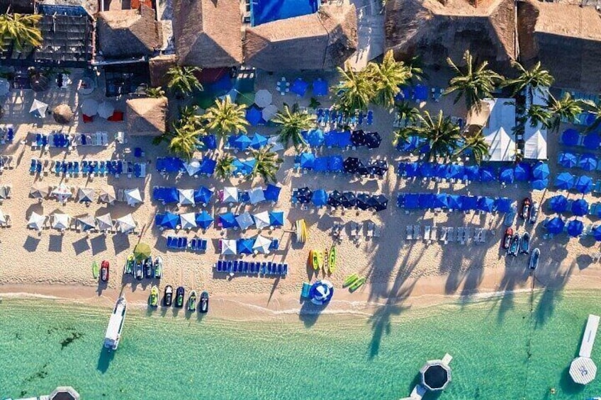 An aerial view of a beach with rows of blue umbrellas and lounge chairs, palm trees, and a few boats. 2