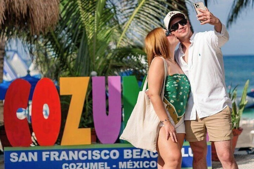 A couple taking a selfie in front of a colorful sign with the text 'COZUMEL' and 'SAN FRANCISCO BEACH COZUMEL - MÉXICO'. 3