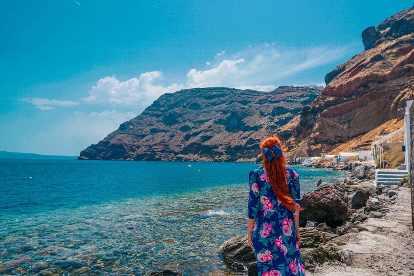 A woman in a floral dress standing by a rocky shoreline with clear blue water and a mountainous backdrop. 4