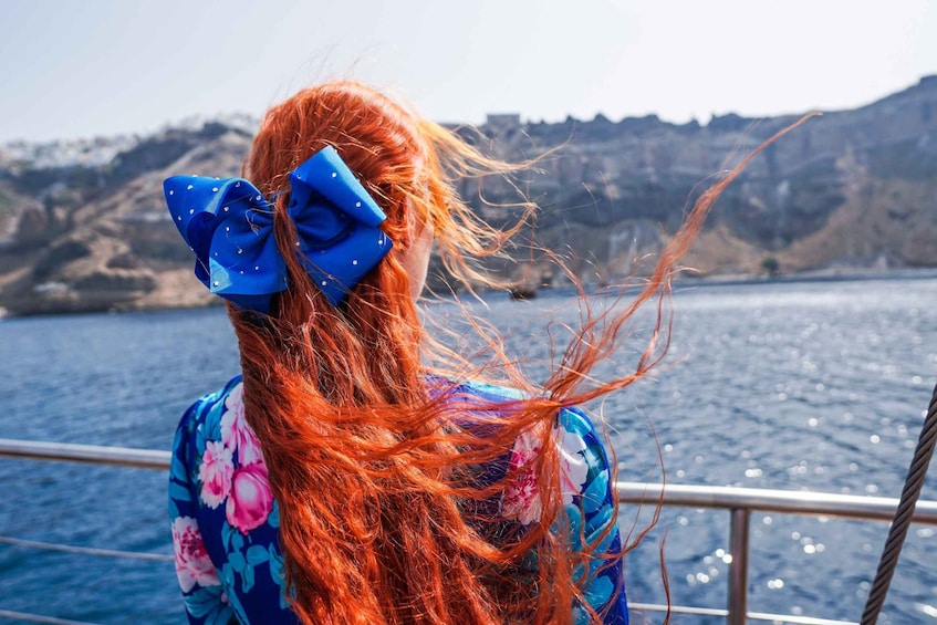 A person with long red hair tied with a blue bow, wearing a floral dress, overlooking a body of water with rocky hills in the background. 2