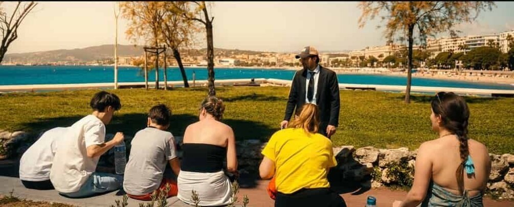 A group of people sitting on a grassy area by a waterfront, with a man standing in the center. 5