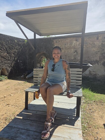 A person sitting on a wooden bench outdoors with a stone wall and a metal structure in the background. 2