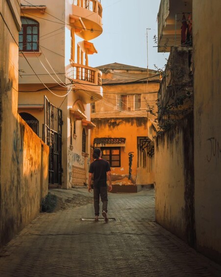 A narrow alleyway with a person walking away from the camera, buildings with balconies, and a door with graffiti. 5