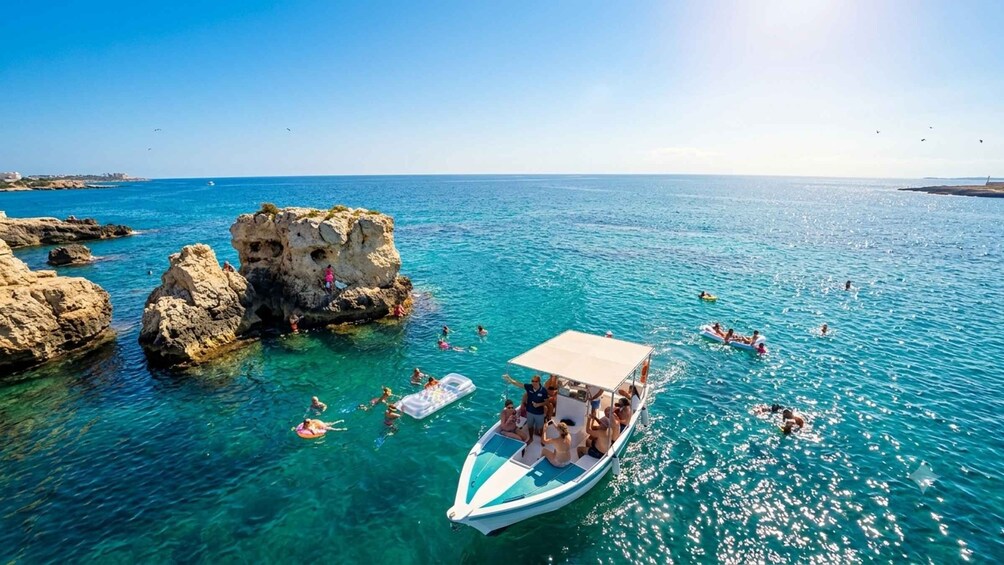 A clear blue sea with rocky outcrops, a boat with people on board, and individuals swimming and floating. 1