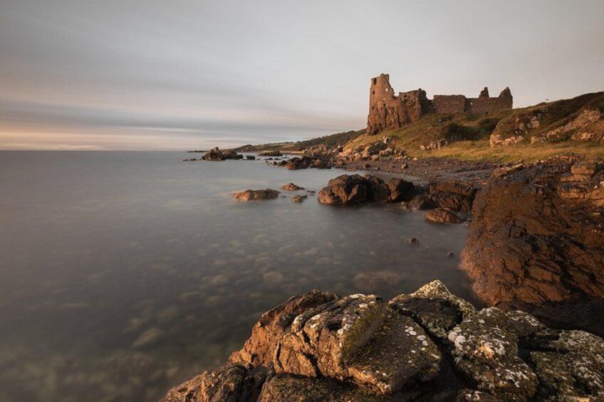 A coastal landscape with a ruined castle, rocky shore, and calm sea. 2