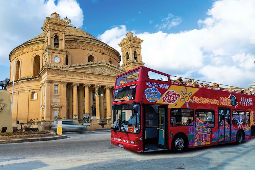 A red double-decker bus with "CitySightseeing Malta" written on it, parked in front of a historic building with a dome and columns. 1