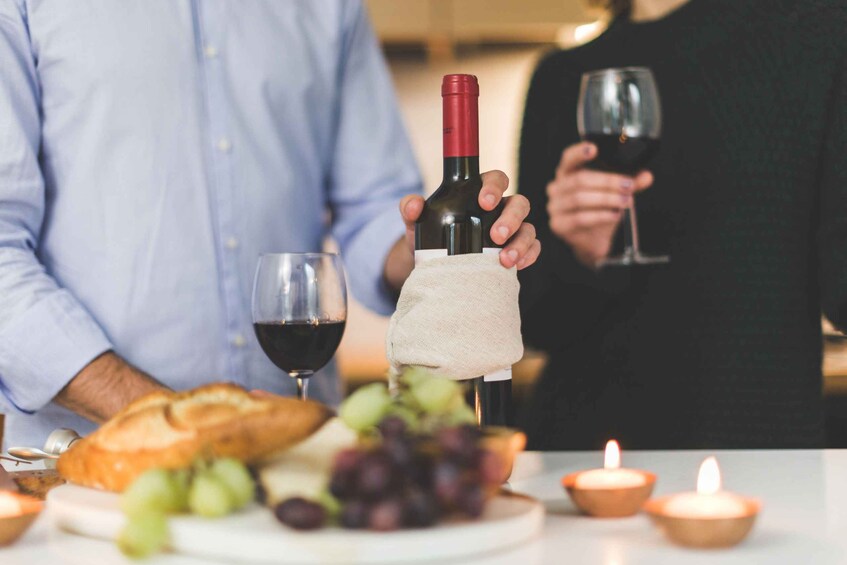 Una persona sosteniendo una botella de vino con un paño, una copa de vino, pan, uvas y velas encendidas sobre una mesa. 1