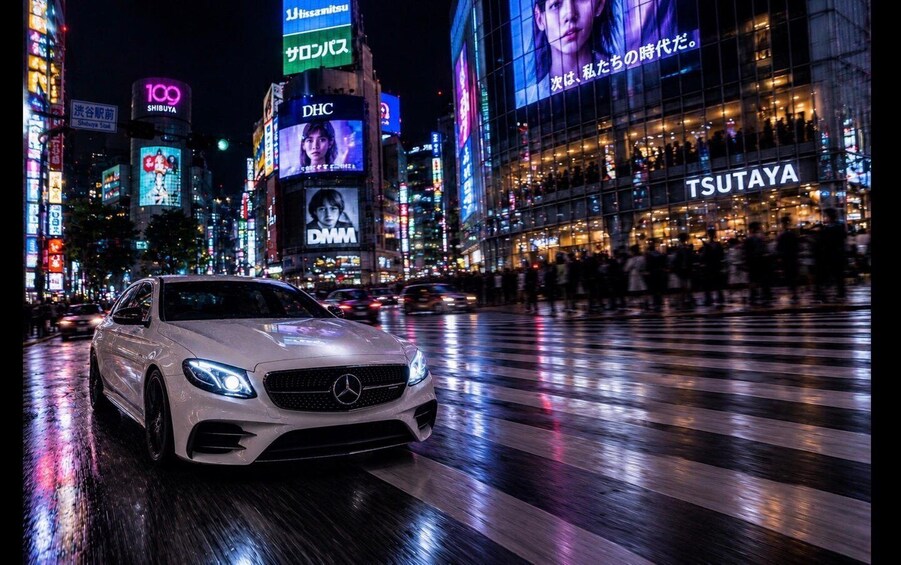 A white car on a wet street with illuminated billboards and a building labeled 'TSUTAYA' in the background. 1