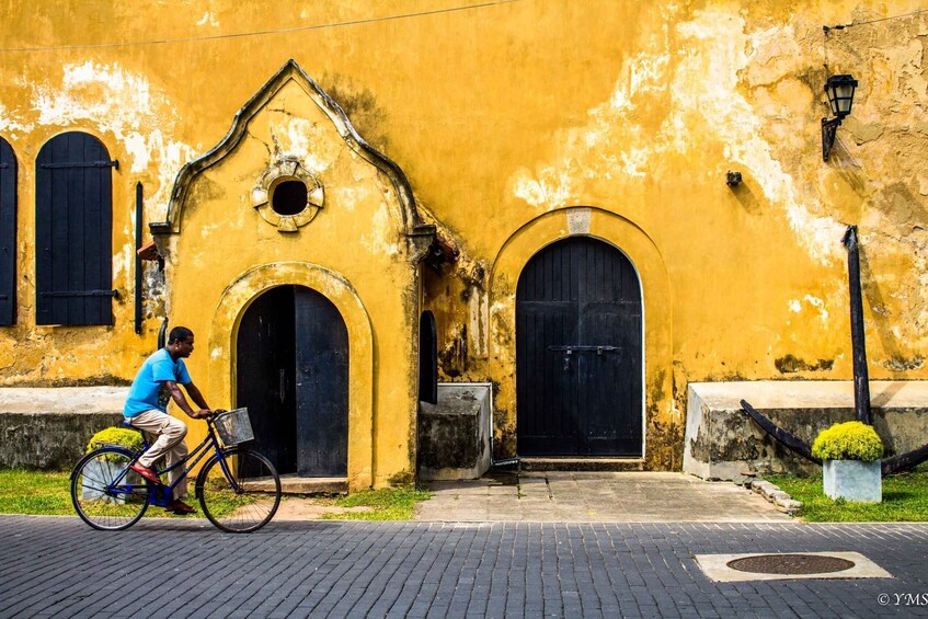 A man riding a bicycle past a yellow building with arched windows and doors. 1