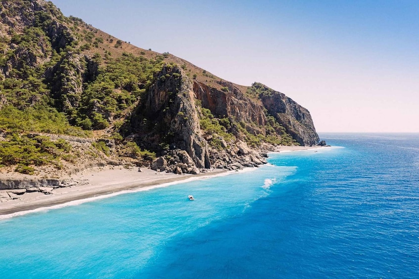 A coastal landscape with a clear blue sea, a sandy beach, and a rocky cliff with sparse vegetation. 5