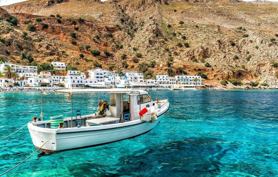 A boat on clear turquoise water with white buildings and a rocky hillside in the background. 3