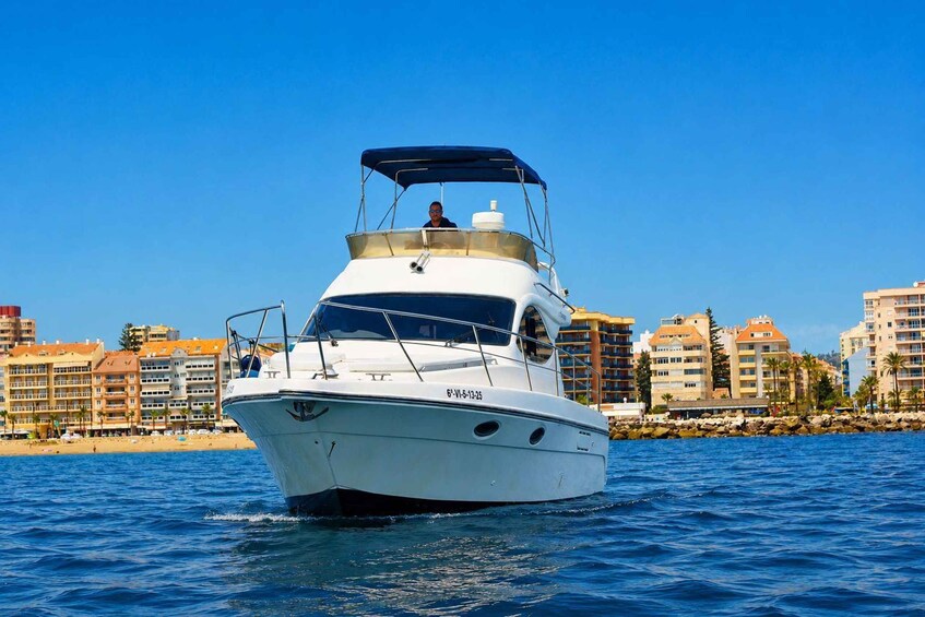 A white yacht with a blue canopy on the water, with a coastal cityscape in the background. 2
