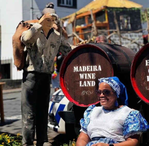 A person sitting in front of barrels labeled 'MADEIRA WINE LAND' with a statue of a person in the background. 1
