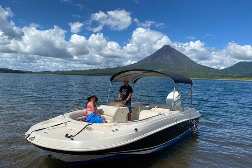 A motorboat on calm water with a person steering and another person seated, a mountain in the background, and a clear sky. 3