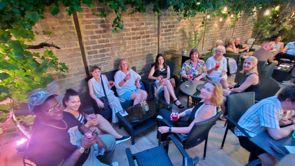 A group of people sitting outdoors at a social gathering, with a brick wall and greenery in the background. 3